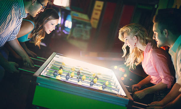Two couples playing foosbal aginst each other on weekend night. They are in mid 20's. People are lit by light from foosball table and very little to zero ambiental light. Tilt shot,side view.